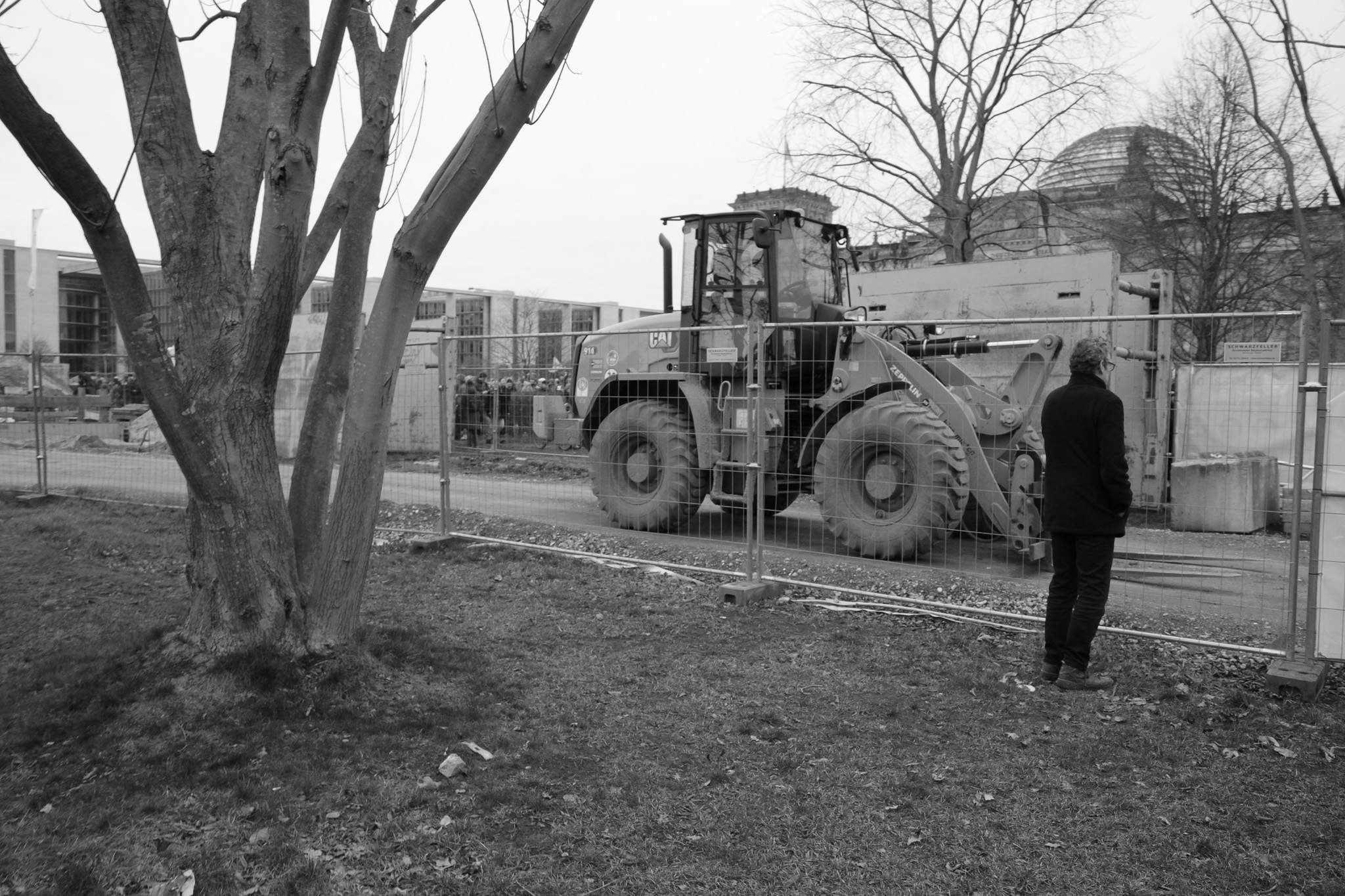 Person standing before a wheel loader behind construction fencing, black and white