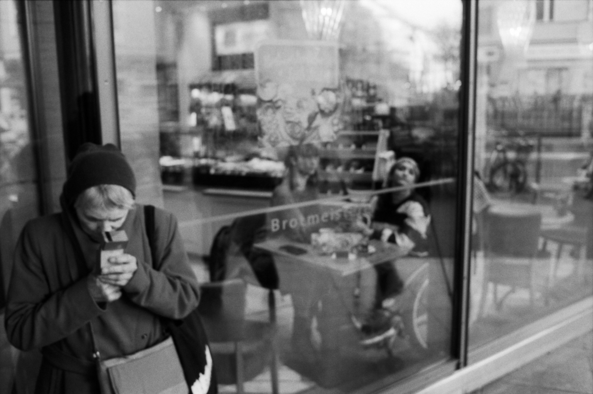 Person checking their phone outside a bakery window with reflections, black and white