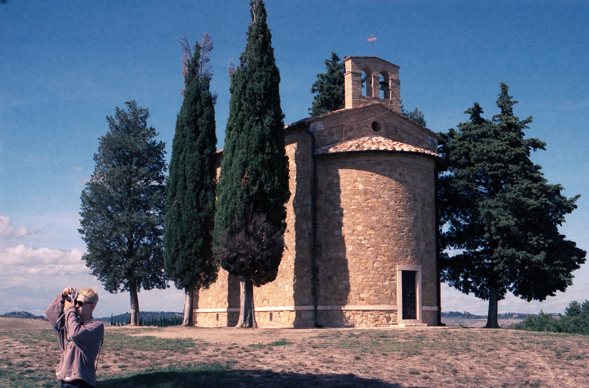 Person photographing a stone chapel with cypress trees, Tuscany