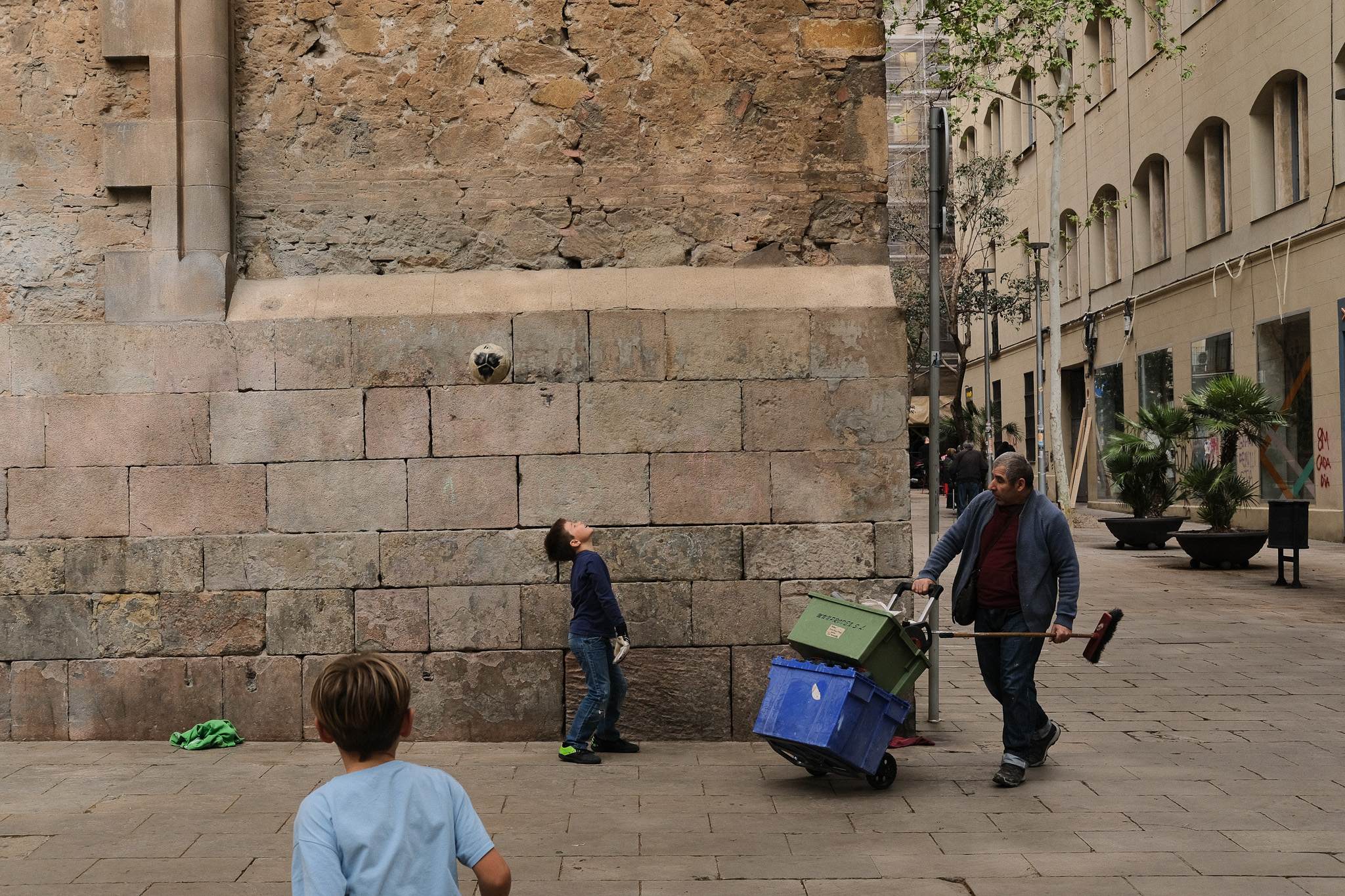 Child looking up at an ancient stone wall while a man pushes a cart, Barcelona